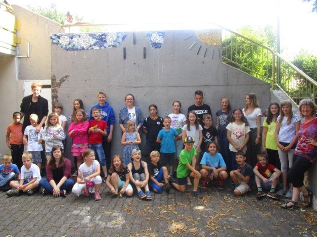 Große Gruppe von Kindern und Erwachsenen vor einer Mosaik-Sonnenwand.